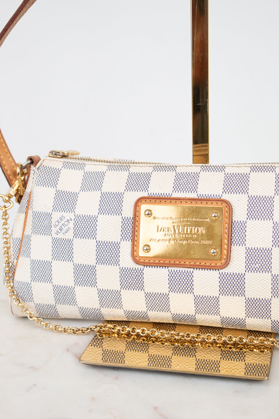 A Louis Vuitton shoulder bag in Damier Azur canvas with a blue and white checkered pattern, tan leather trim, and a gold-tone metal plaque and on a light marble background.