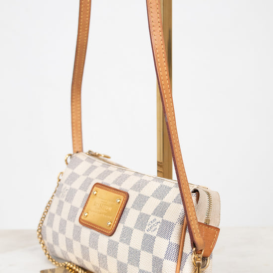 A small crossbody handbag with a gray and white checkerboard pattern, tan leather trim, and a gold-tone chain strap, displayed on a stand against a light background.