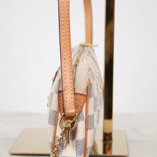 A close-up side view of a Louis Vuitton shoulder bag in Damier Ebene canvas with tan leather trim, resting on a gold-colored display stand.