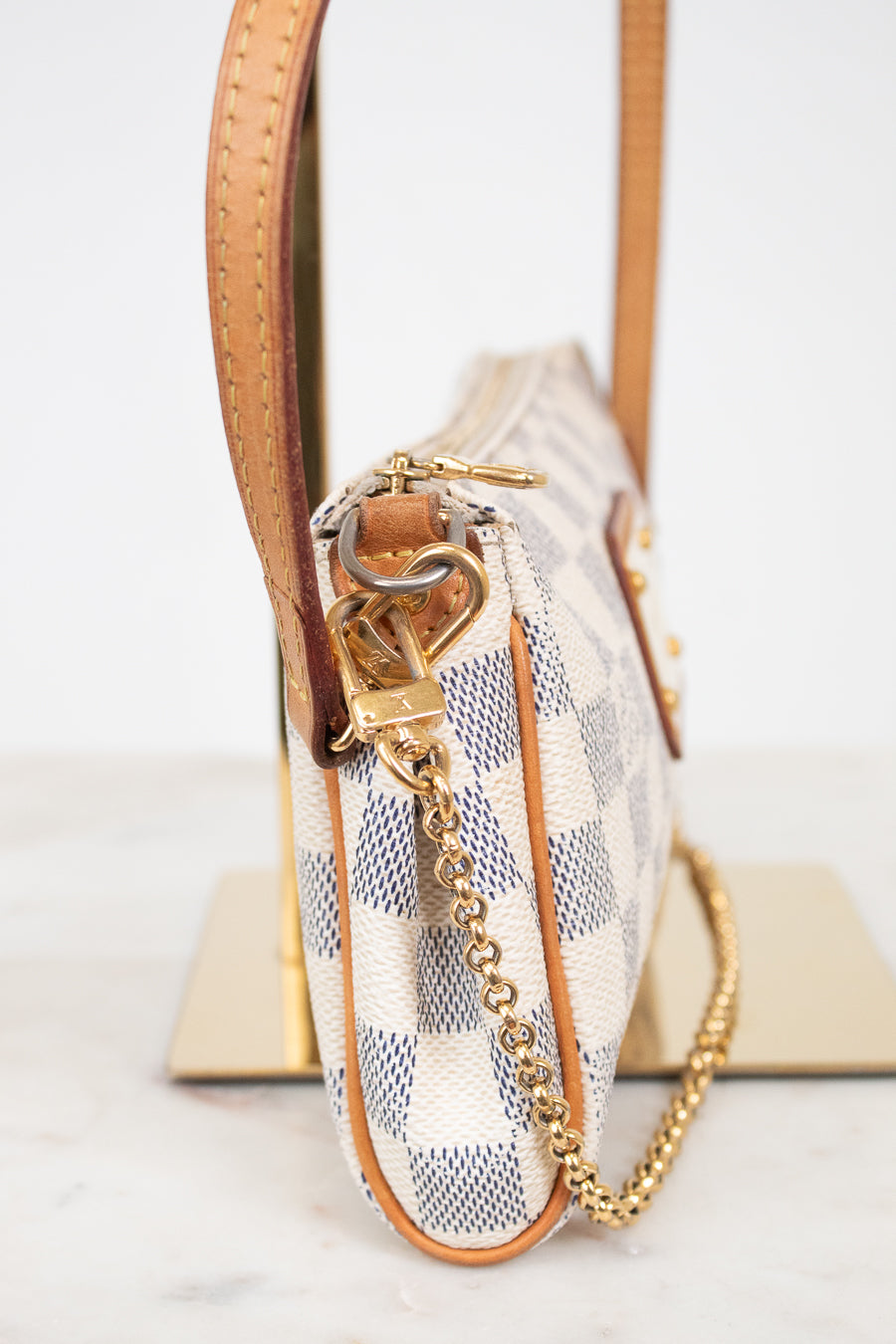 A close-up side view of a Louis Vuitton shoulder bag in Damier Azur canvas, featuring a beige and blue checkered pattern, tan leather trim, and a gold-tone chain strap.