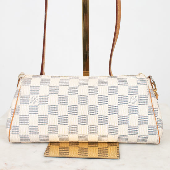 A Louis Vuitton shoulder bag in Damier Azur coated canvas, featuring a cream and light gray checkered pattern with tan leather trim and gold-tone hardware, displayed on a stand.