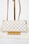 A Louis Vuitton shoulder bag in Damier Azur coated canvas, featuring a cream and light gray checkered pattern with tan leather trim and gold-tone hardware, displayed on a stand.
