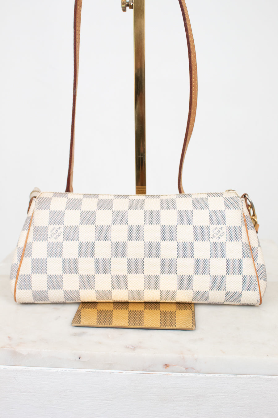 A Louis Vuitton shoulder bag in Damier Azur coated canvas, featuring a cream and light gray checkered pattern with tan leather trim and gold-tone hardware, displayed on a stand.