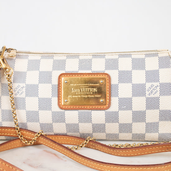 A Louis Vuitton shoulder bag in the Damier Azur coated canvas, featuring a blue and white checkered pattern, tan leather trim, a gold-tone metal plaque, and a gold chain strap, displayed on a light marble surface against a white background.