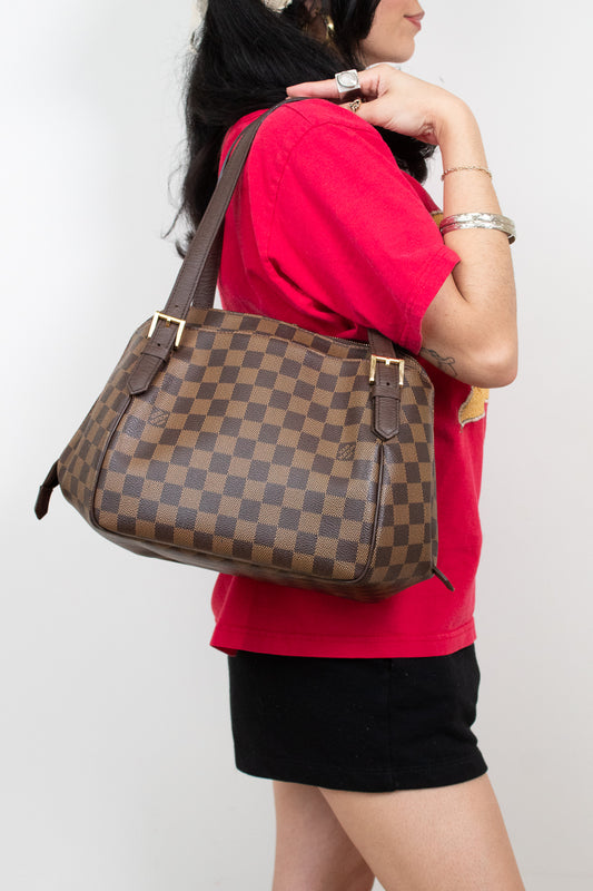 A woman wearing a red t-shirt and black shorts carries a brown checkered Louis Vuitton shoulder bag against a plain white background.