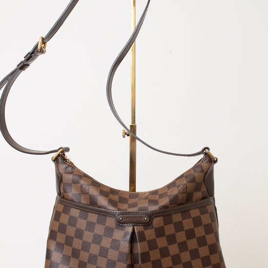 A brown and dark brown checkered shoulder bag with a long strap, displayed on a gold stand against a plain white background.