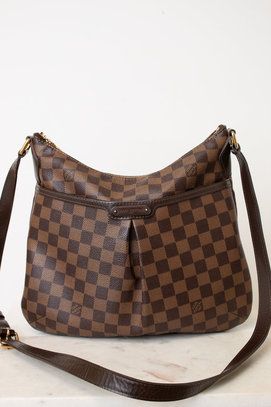A brown checkered shoulder bag with a dark brown leather strap, resting on a light marble surface against a plain white background.