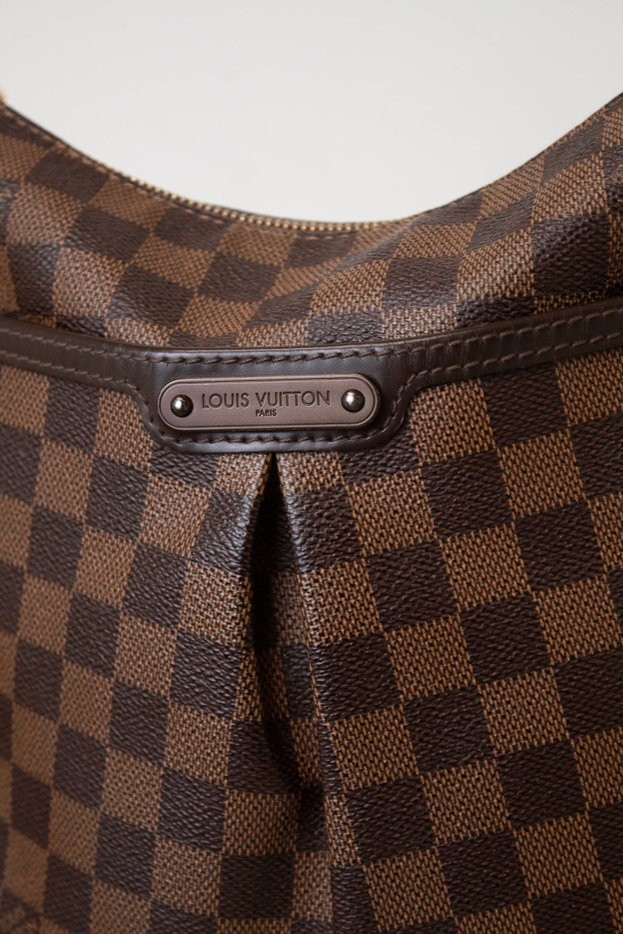 Close-up of a Louis Vuitton handbag in the Damier Ebene check pattern, showing the brown and tan checkerboard canvas and a metal brand plaque.