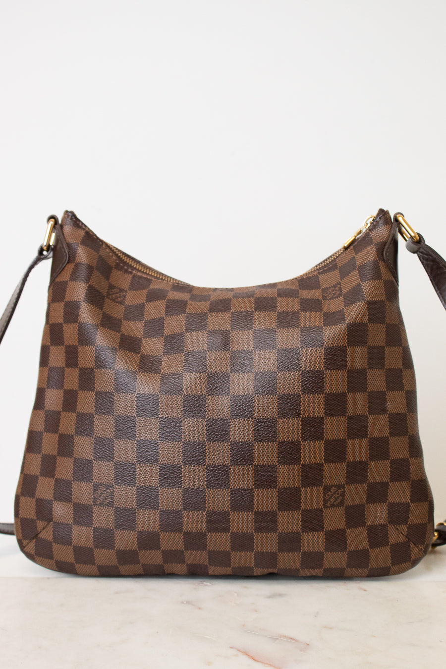 A brown and tan checkered shoulder bag with gold-tone hardware, resting on a light marble surface against a plain white background.