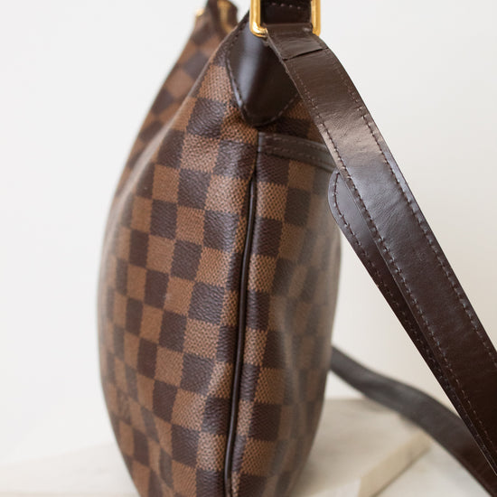 A brown checkered shoulder bag with a dark brown leather strap and gold-tone hardware, displayed on a white marble block against a white background.