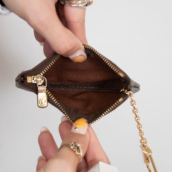 Brown pouch with gold zipper and chain held by a hand against a light gray background