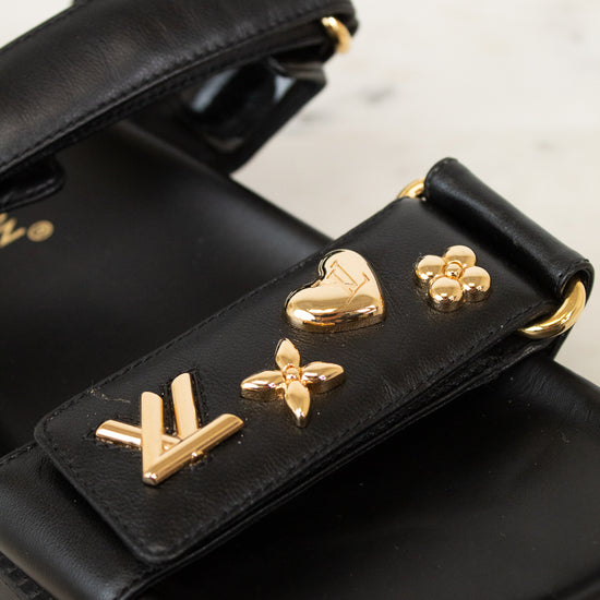 A close-up of a black leather sandal featuring gold-tone Louis Vuitton-inspired monogram and floral charms on the strap, set against a light marble background.