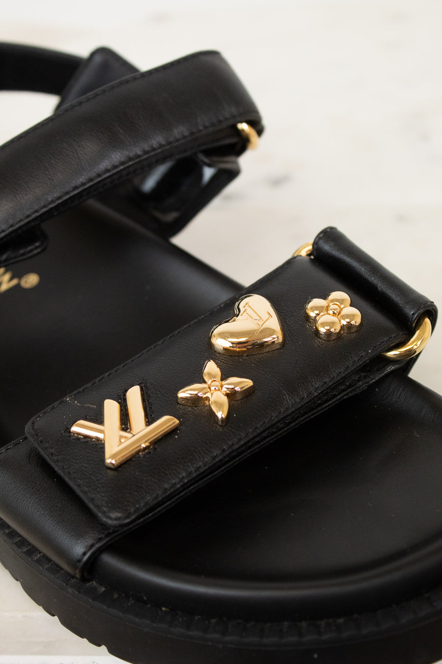 A close-up of a black leather sandal featuring gold-tone Louis Vuitton-inspired monogram and floral charms on the strap, set against a light marble background.