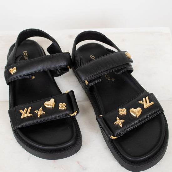 A pair of black leather sandals with gold-tone Louis Vuitton monogram and heart charms, displayed on a light marble surface.