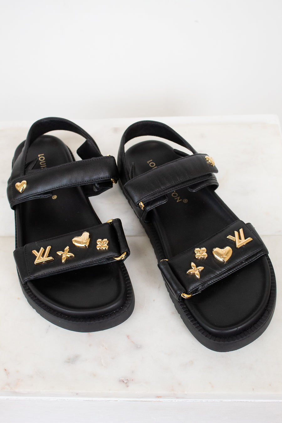 A pair of black leather sandals with gold-tone Louis Vuitton monogram and heart charms, displayed on a light marble surface.