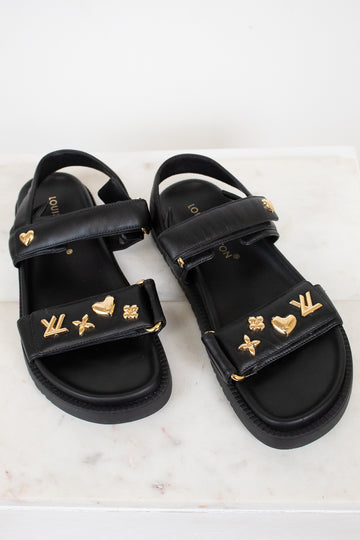 A pair of black leather sandals with gold-tone Louis Vuitton monogram and heart charms, displayed on a light marble surface.