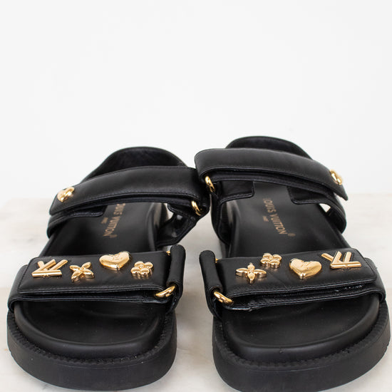 A pair of black sandals with multiple wide straps adorned with gold-tone metal charms, including LV monogram, hearts, stars, and floral motifs, displayed on a light marble surface against a white background.