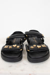A pair of black sandals with multiple wide straps adorned with gold-tone metal charms, including LV monogram, hearts, stars, and floral motifs, displayed on a light marble surface against a white background.