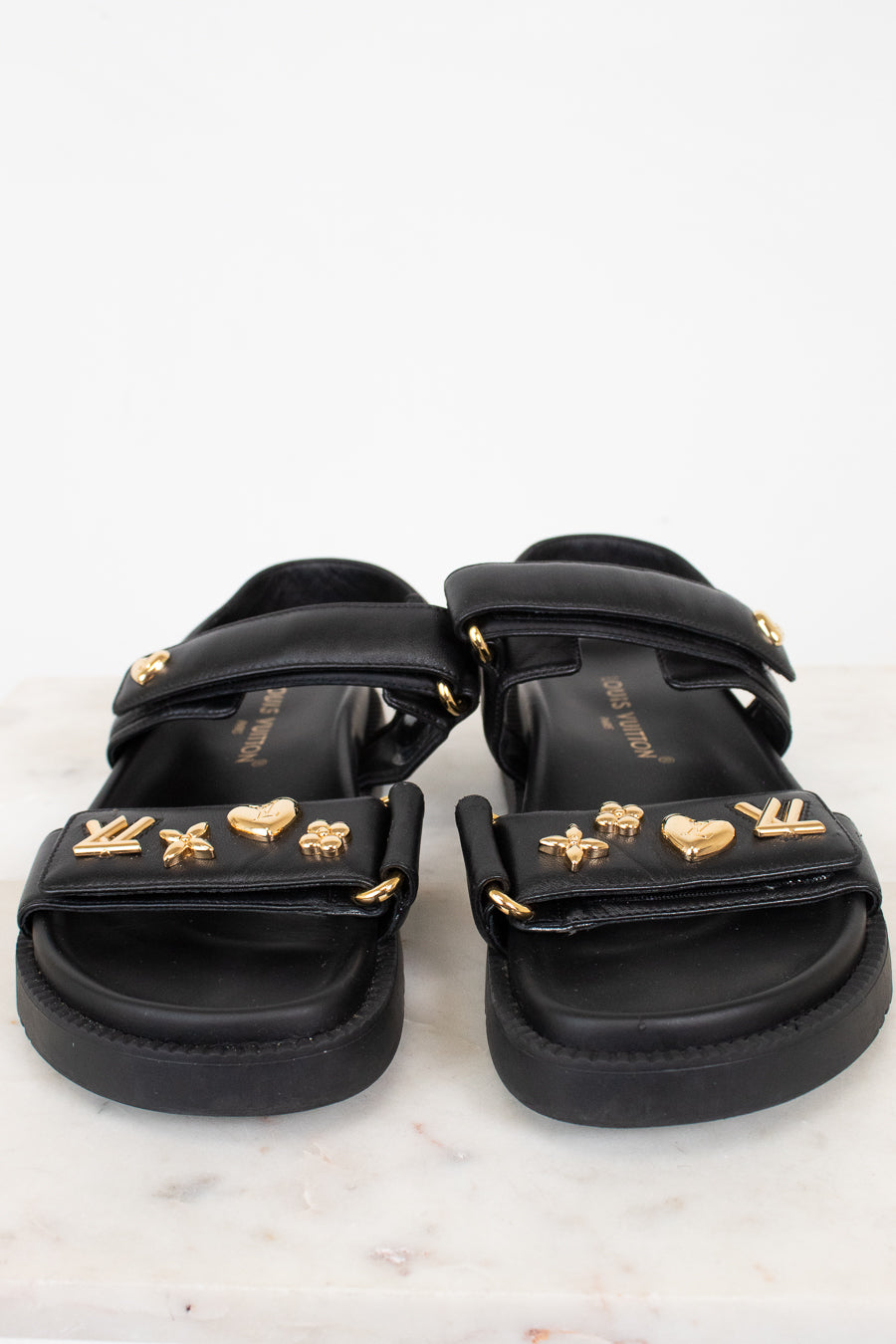 A pair of black sandals with multiple wide straps adorned with gold-tone metal charms, including LV monogram, hearts, stars, and floral motifs, displayed on a light marble surface against a white background.