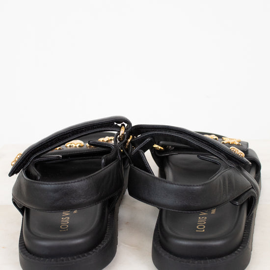A pair of black leather platform sandals with gold-tone hardware and straps, shown from a rear angle on a light marble surface against a white background.