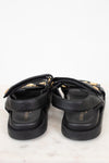 A pair of black leather platform sandals with gold-tone hardware and straps, shown from a rear angle on a light marble surface against a white background.