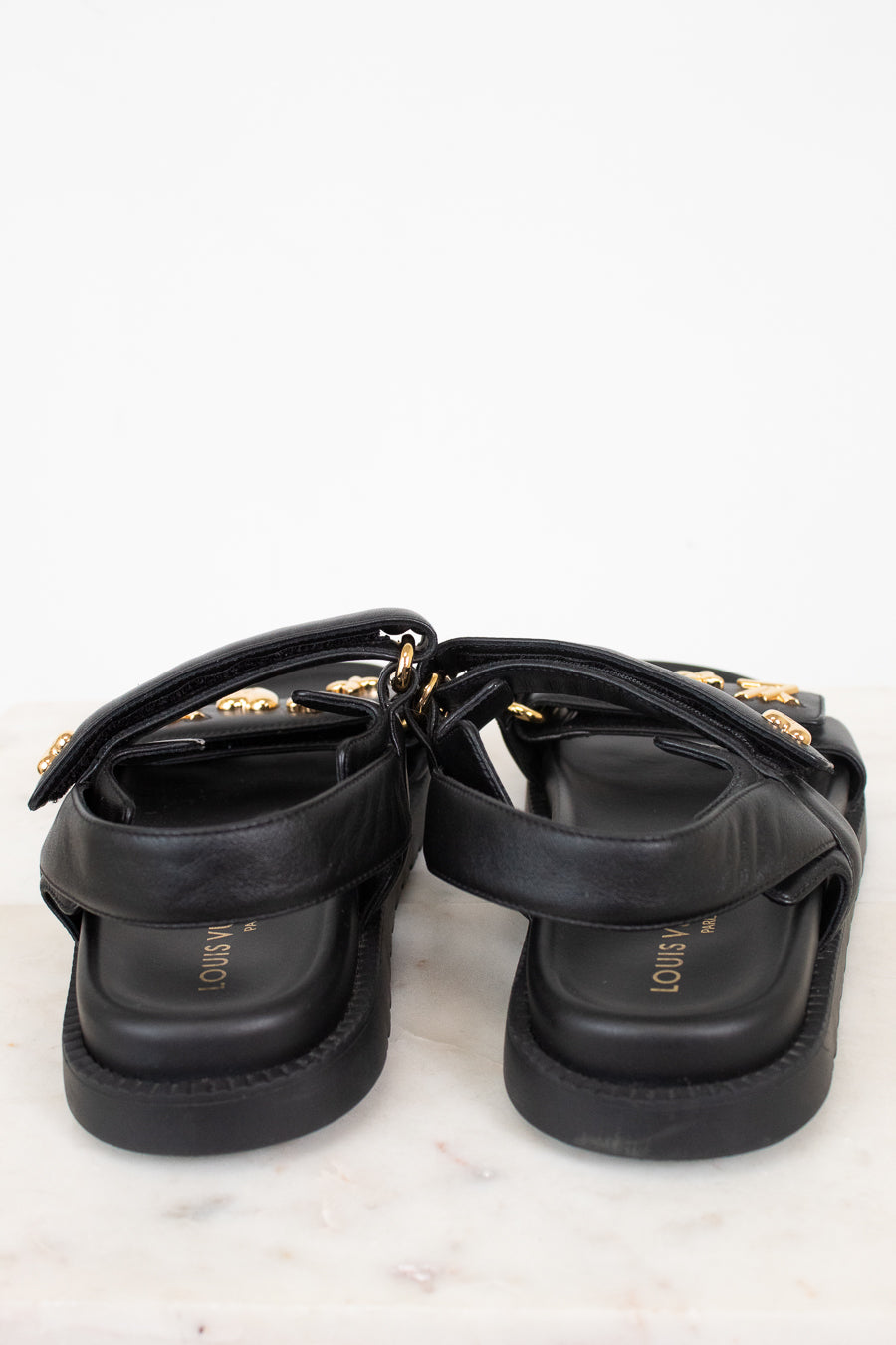 A pair of black leather platform sandals with gold-tone hardware and straps, shown from a rear angle on a light marble surface against a white background.