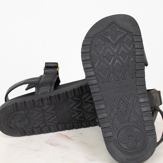 A pair of black leather sandals shown from the sole side, resting on a white marble surface against a white background.