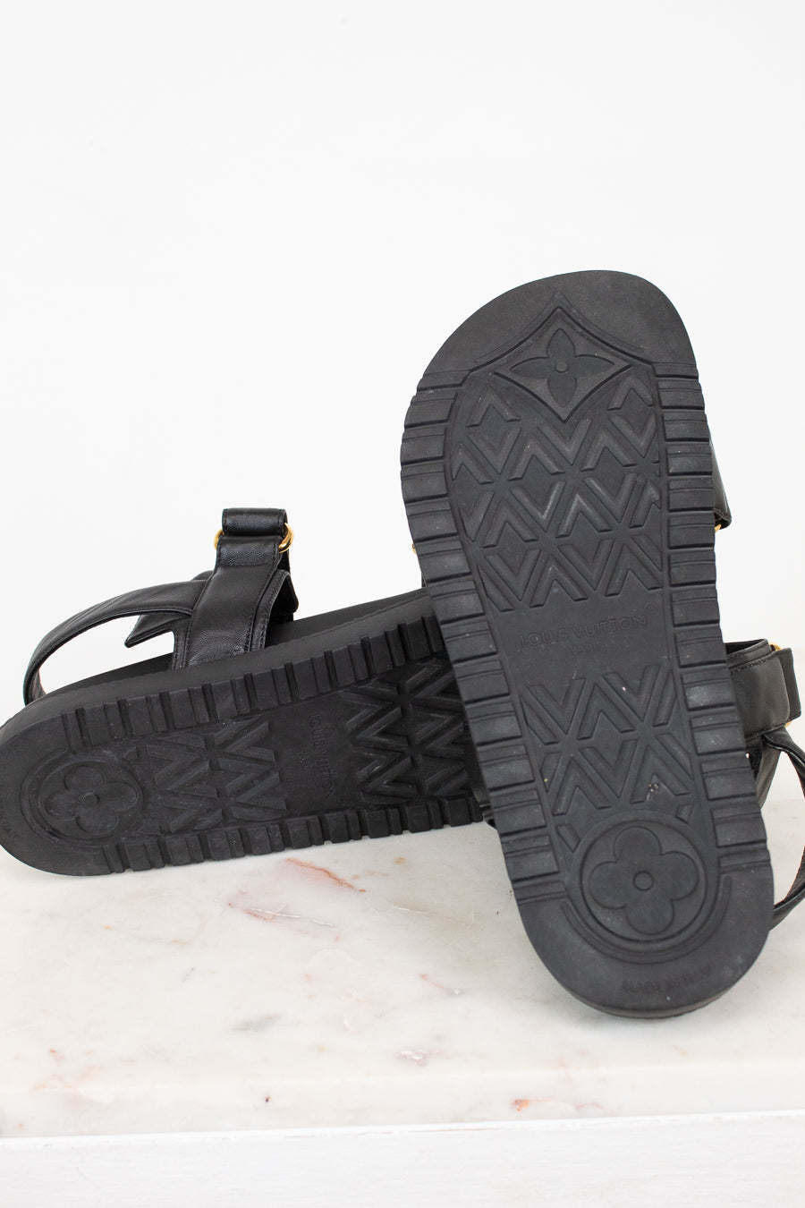A pair of black leather sandals shown from the sole side, resting on a white marble surface against a white background.