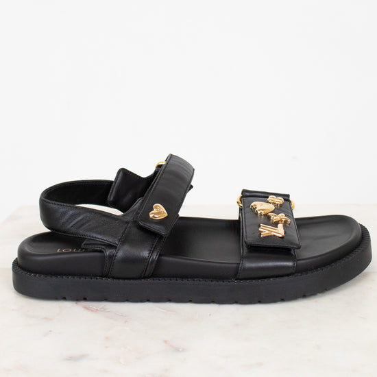 A black leather double-strap sandal with gold-tone LV monogram and heart details, displayed on a white marble surface against a white background.