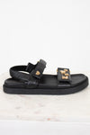 A black leather double-strap sandal with gold-tone LV monogram and heart details, displayed on a white marble surface against a white background.