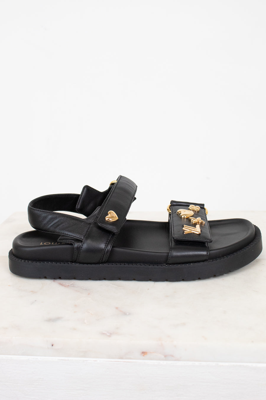 A black leather double-strap sandal with gold-tone LV monogram and heart details, displayed on a white marble surface against a white background.
