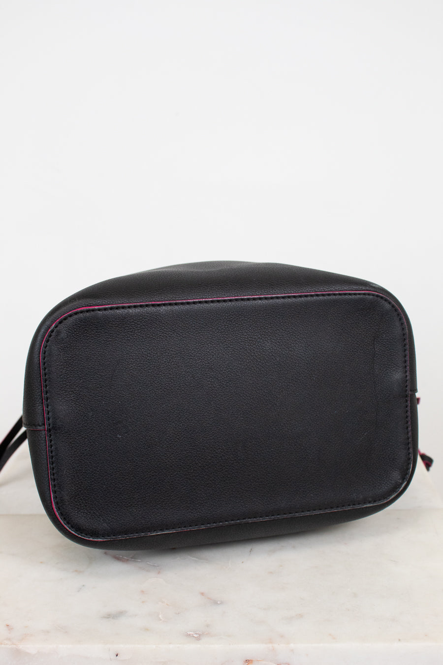 Bottom view of black leather handbag with rounded shape bottom and contrasting pink stitching, resting on a light marble surface against a white background.