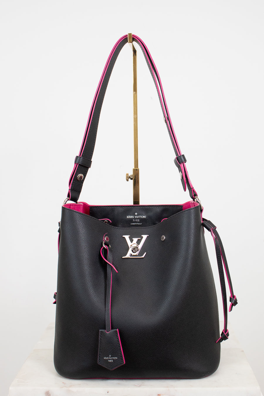 A black leather Louis Vuitton bucket bag with vibrant pink lining and trim, featuring a silver-tone LV logo on the front and a hanging leather tag, displayed on a stand against a white background.