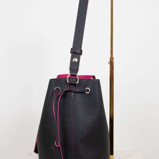 Black leather bucket bag with bright pink contrast stitching and drawstring, displayed on a white marble surface next to a gold metal stand.