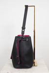 Black leather bucket bag with bright pink contrast stitching and drawstring, displayed on a white marble surface next to a gold metal stand.