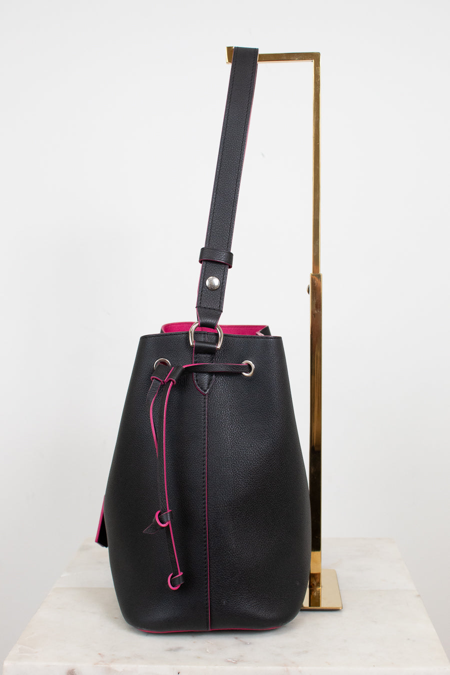 Black leather bucket bag with bright pink contrast stitching and drawstring, displayed on a white marble surface next to a gold metal stand.
