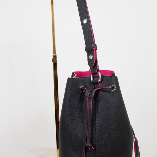 Black leather bucket bag with a vibrant pink interior lining and drawstring closure, displayed on a gold stand against a white background.