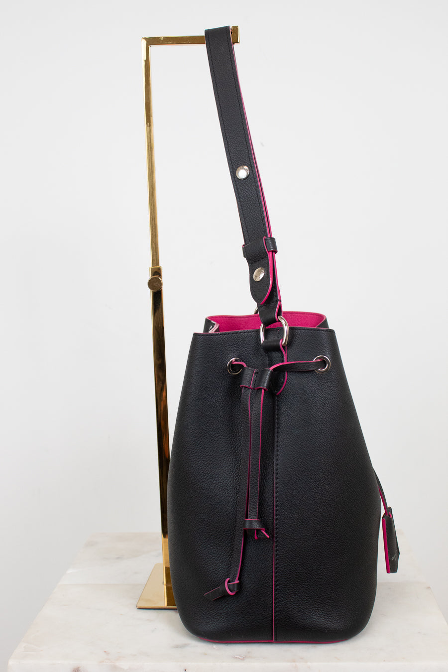 Black leather bucket bag with a vibrant pink interior lining and drawstring closure, displayed on a gold stand against a white background.