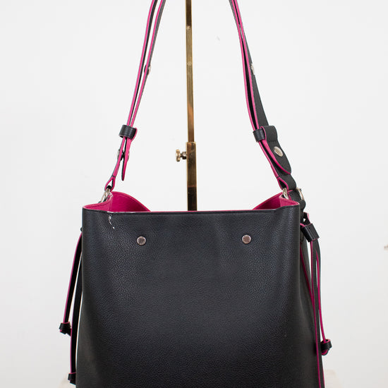 A black leather bucket tote bag with a vibrant pink interior lining and matching shoulder straps, displayed on a stand against a white background.