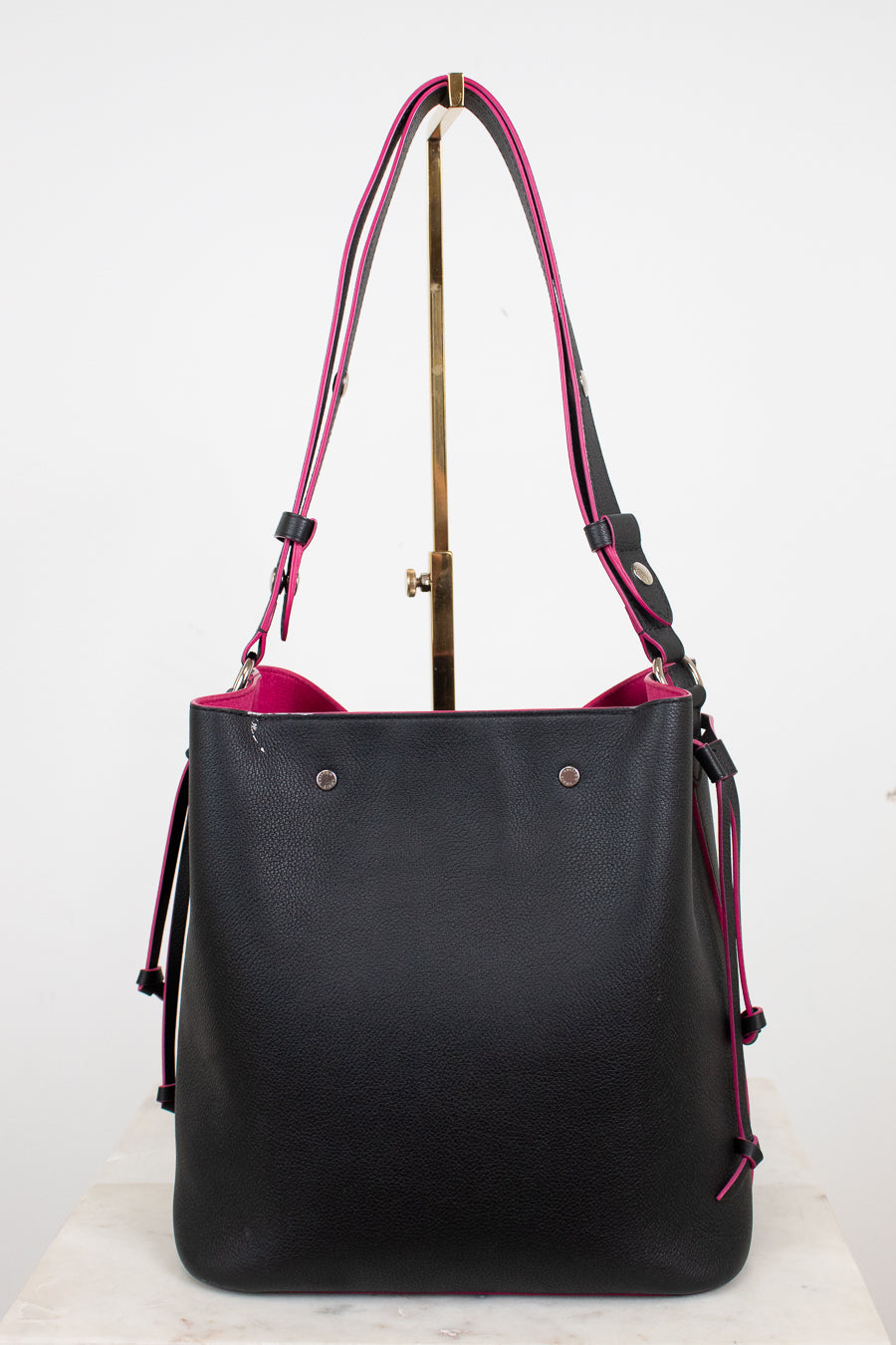 A black leather bucket tote bag with a vibrant pink interior lining and matching shoulder straps, displayed on a stand against a white background.