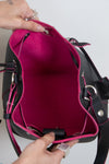 A close-up of a black leather handbag with a bright pink interior lining, held open by two hands against a plain light background.