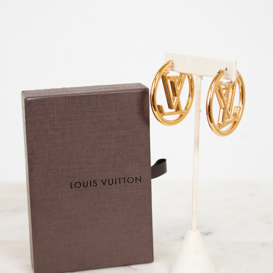 Gold earrings on a stand next to a Louis Vuitton box on a white background