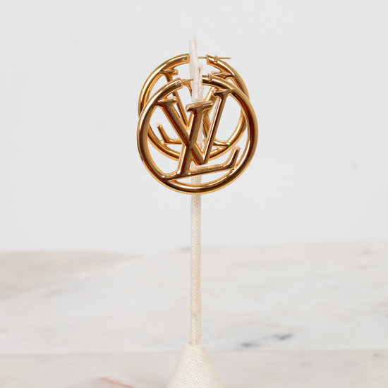Gold circular hoop earrings with a logo on a white stand against a light background
