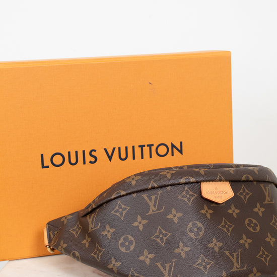 A Louis Vuitton monogram bumbag pouch in brown coated canvas with tan leather accents, shown in front of its signature orange Louis Vuitton box on a white marble surface.