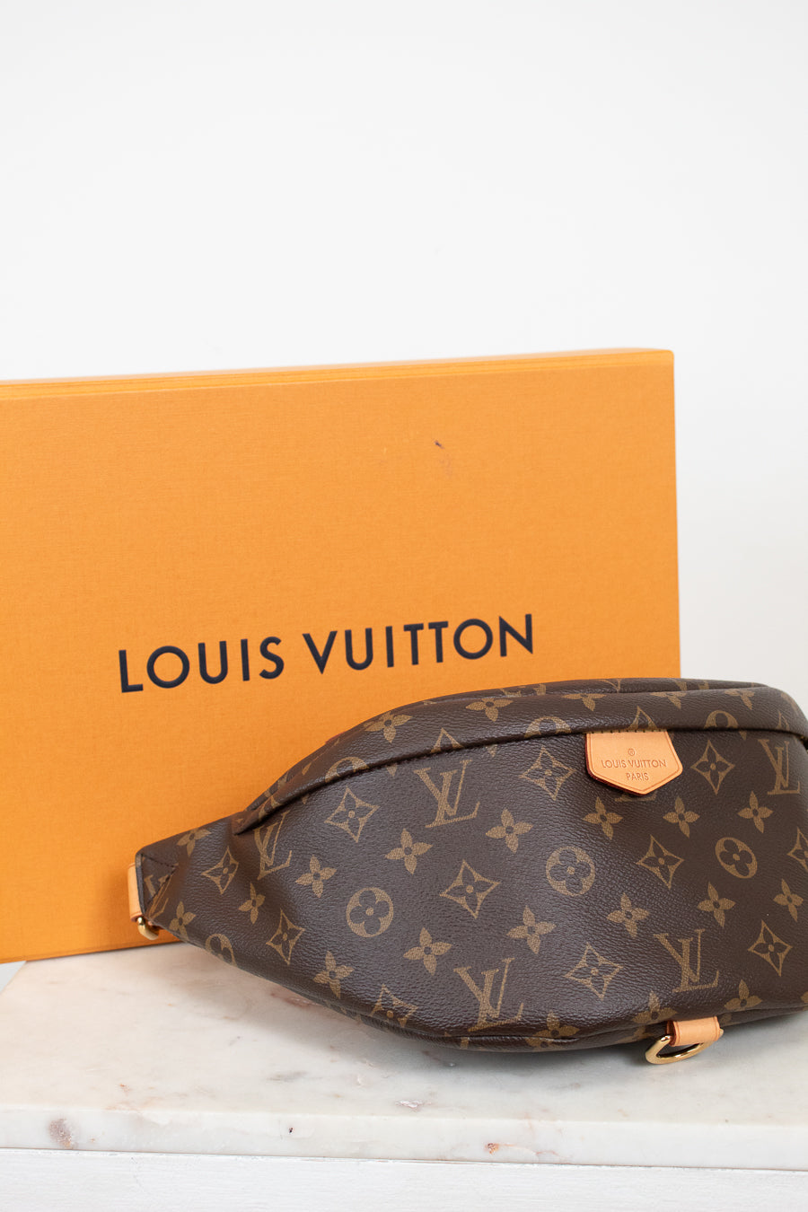 A Louis Vuitton monogram bumbag pouch in brown coated canvas with tan leather accents, shown in front of its signature orange Louis Vuitton box on a white marble surface.