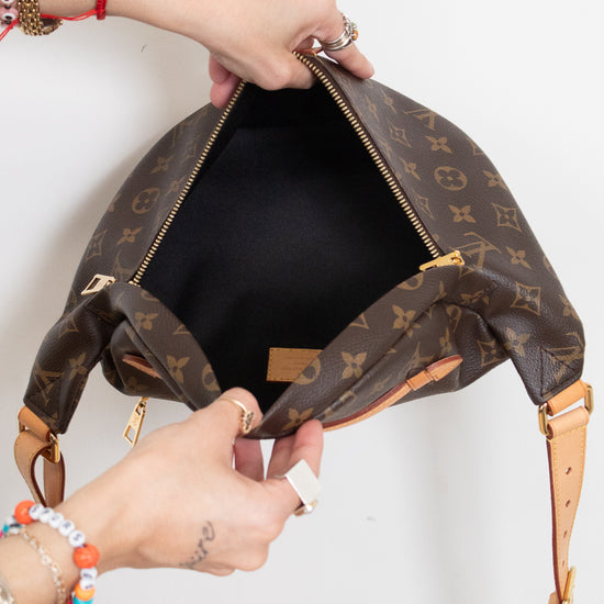 A Louis Vuitton monogram shoulder bag in brown coated canvas with tan leather trim, shown open with a person's hands holding it against a plain white background.