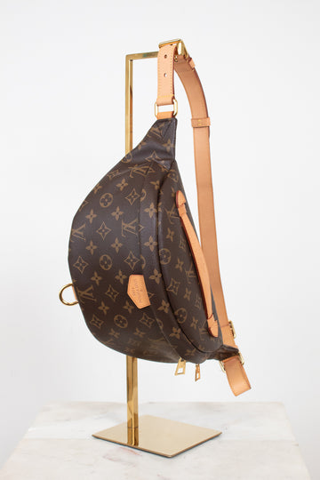 Louis Vuitton monogram bum bag in brown coated canvas with tan leather trim, displayed on a gold metal stand against a white background.