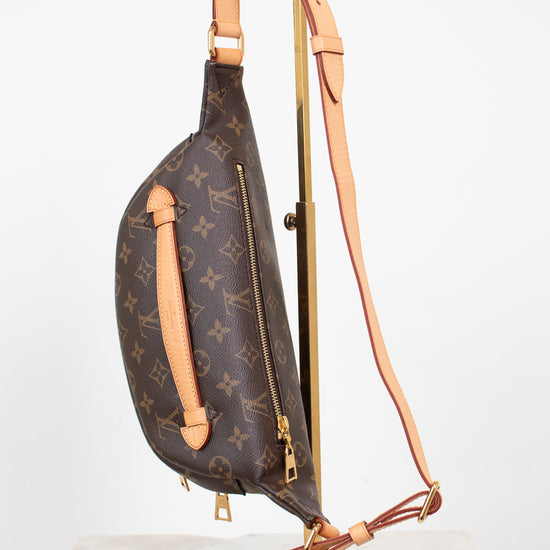 Louis Vuitton monogram canvas sling bag with tan leather trim and a long shoulder strap, displayed on a gold stand against a white background.