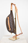 Louis Vuitton monogram canvas sling bag with tan leather trim and a long shoulder strap, displayed on a gold stand against a white background.