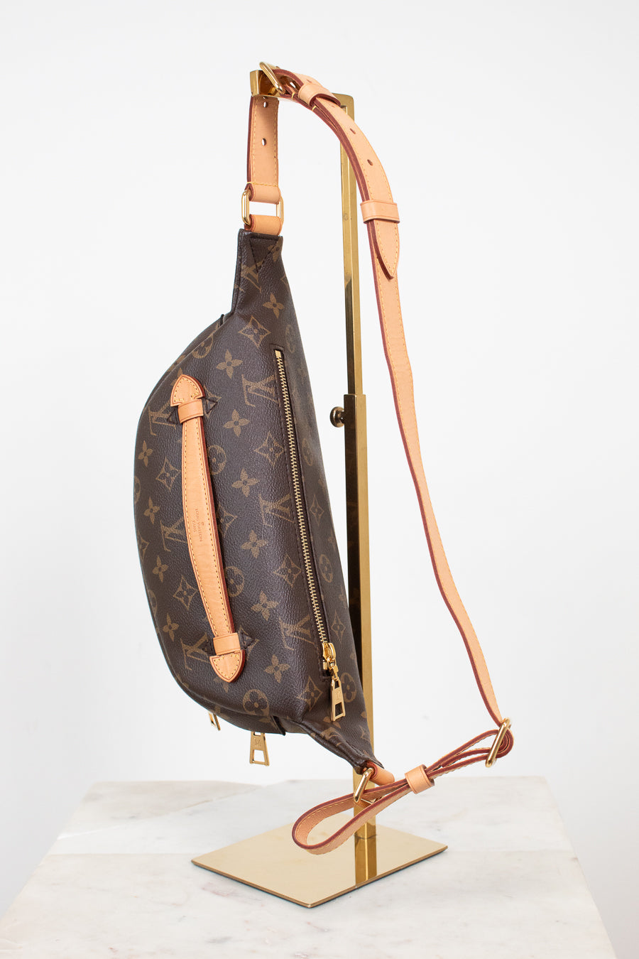 Louis Vuitton monogram canvas sling bag with tan leather trim and a long shoulder strap, displayed on a gold stand against a white background.